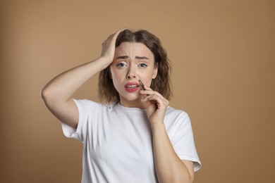 Photo of Disappointed woman plucking her mustache with tweezers on beige background