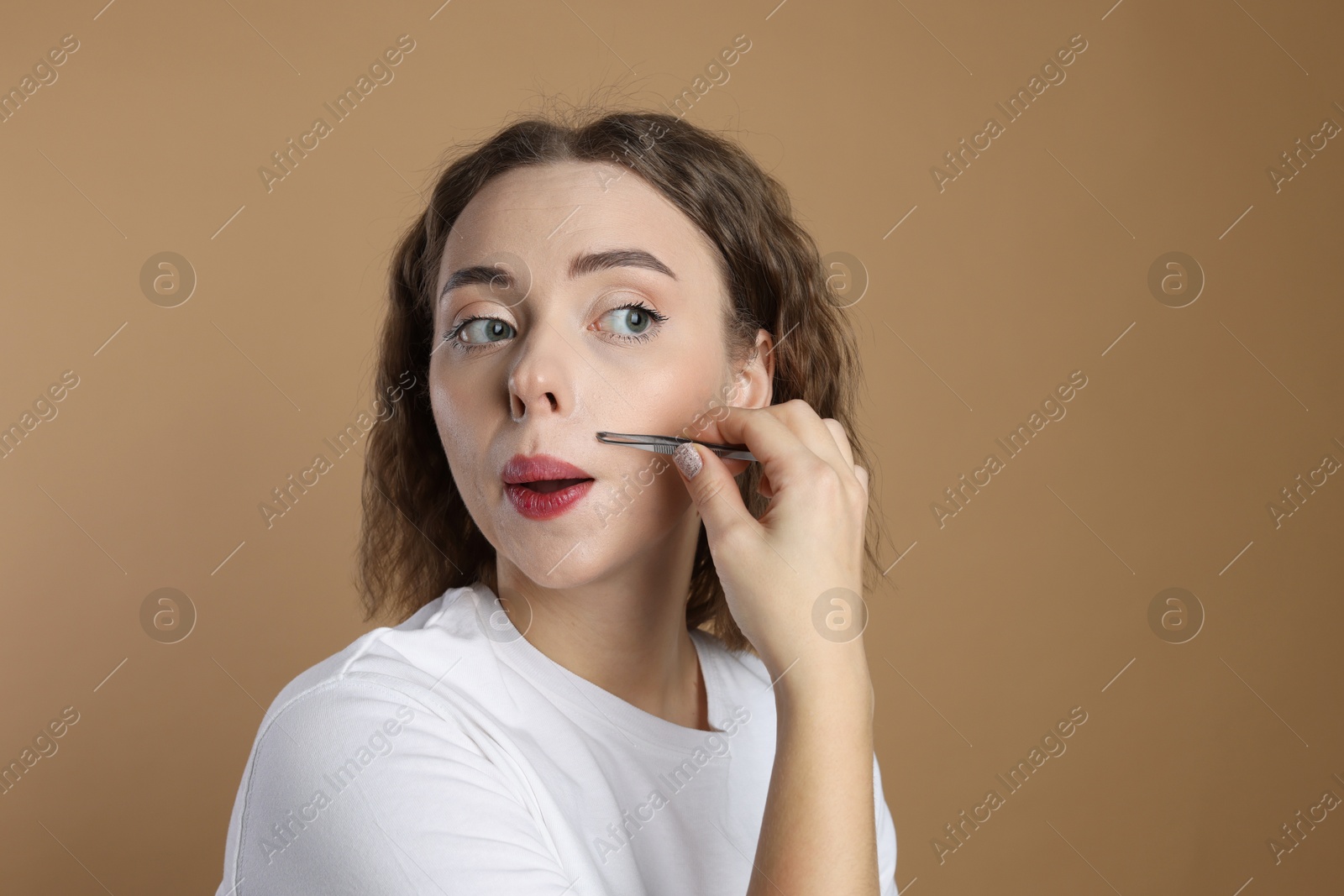 Photo of Beautiful woman plucking her mustache with tweezers on beige background