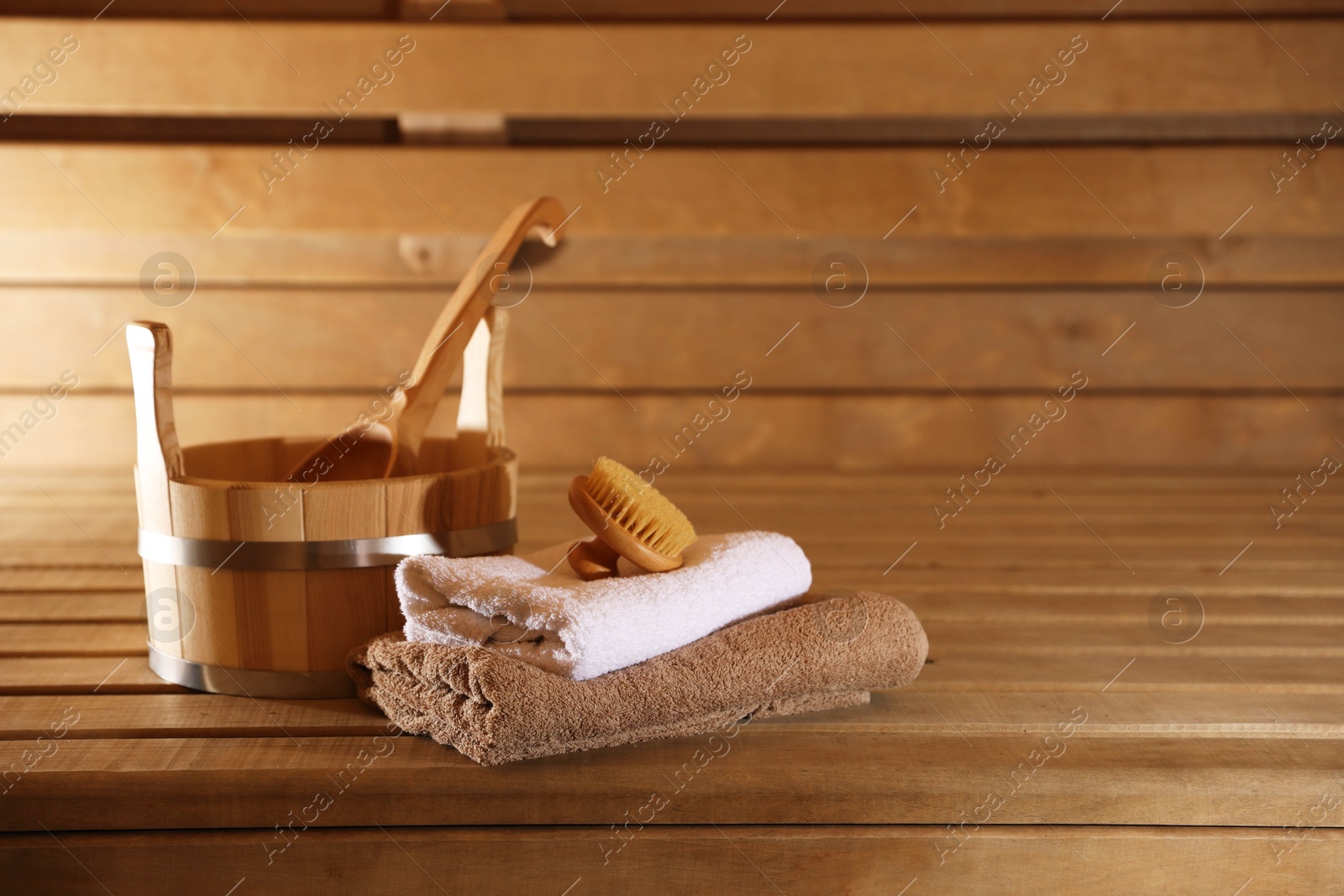 Photo of Bucket, ladle, massaging brush and stack of towels on wooden bench in sauna. Space for text