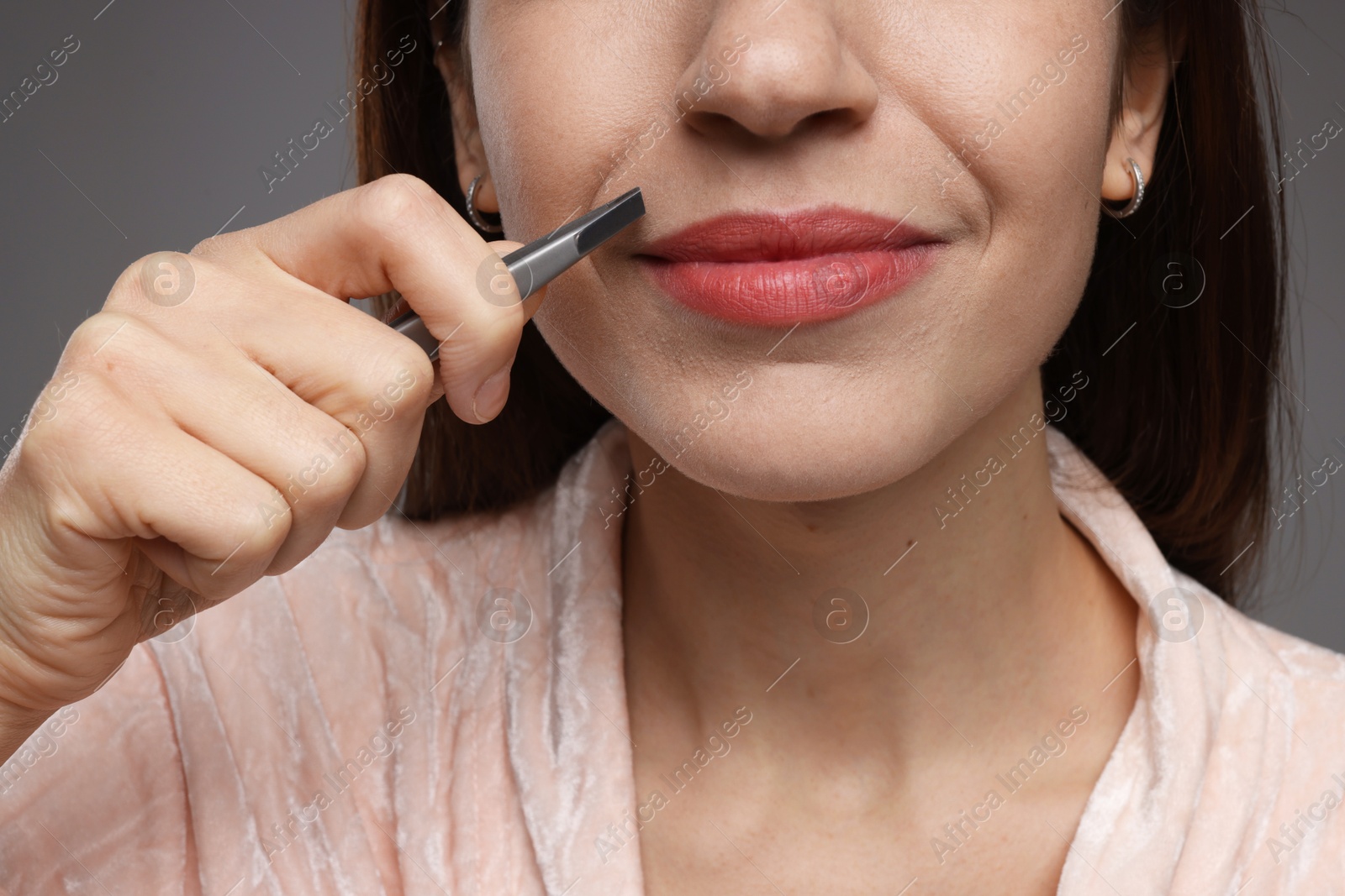 Photo of Woman plucking her mustache with tweezers on grey background, closeup