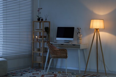 Photo of Comfortable workplace with computer on desk and floor lamp at home office in evening