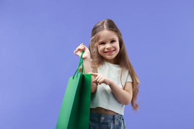 Photo of Happy little girl pointing at shopping bag on violet background