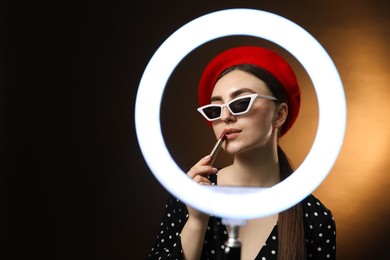 Photo of Beautiful woman applying lipstick on dark background, view through ring lamp. Space for text