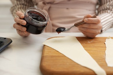 Photo of Woman spreading jam onto fresh dough at white marble table indoors, closeup. Making croissants