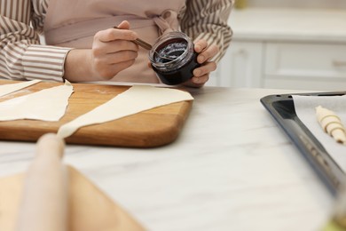 Photo of Woman spreading jam onto fresh dough at white marble table indoors, closeup. Making croissants