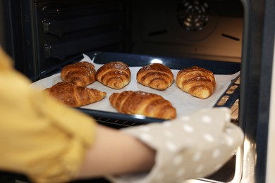 Photo of Woman taking baking sheet with tasty croissants out of oven, closeup