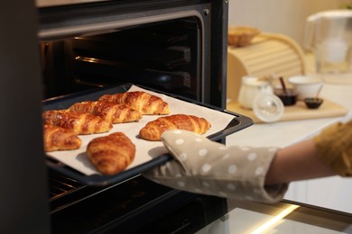 Photo of Woman taking baking sheet with tasty croissants out of oven indoors, closeup