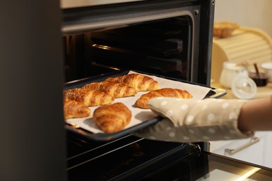 Photo of Woman taking baking sheet with tasty croissants out of oven indoors, closeup