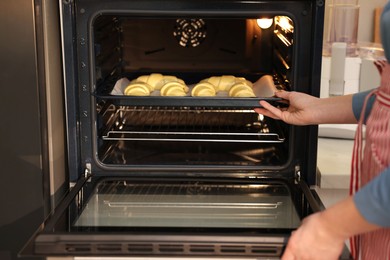 Woman putting baking sheet with raw croissants into oven indoors, closeup