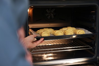 Photo of Woman putting baking sheet with raw croissants into oven, closeup