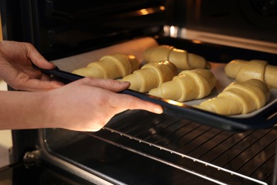 Woman putting baking sheet with raw croissants into oven, closeup