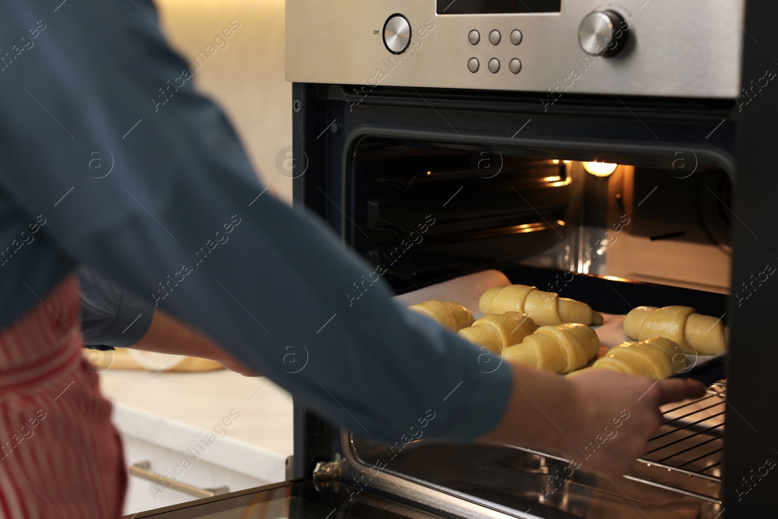 Photo of Woman putting baking sheet with raw croissants into oven indoors, closeup