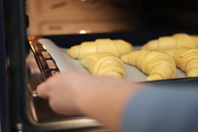 Photo of Woman putting baking sheet with raw croissants into oven, selective focus