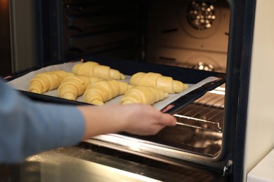 Photo of Woman putting baking sheet with raw croissants into oven, closeup