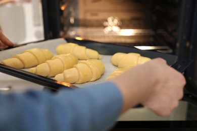 Woman putting baking sheet with raw croissants into oven, closeup