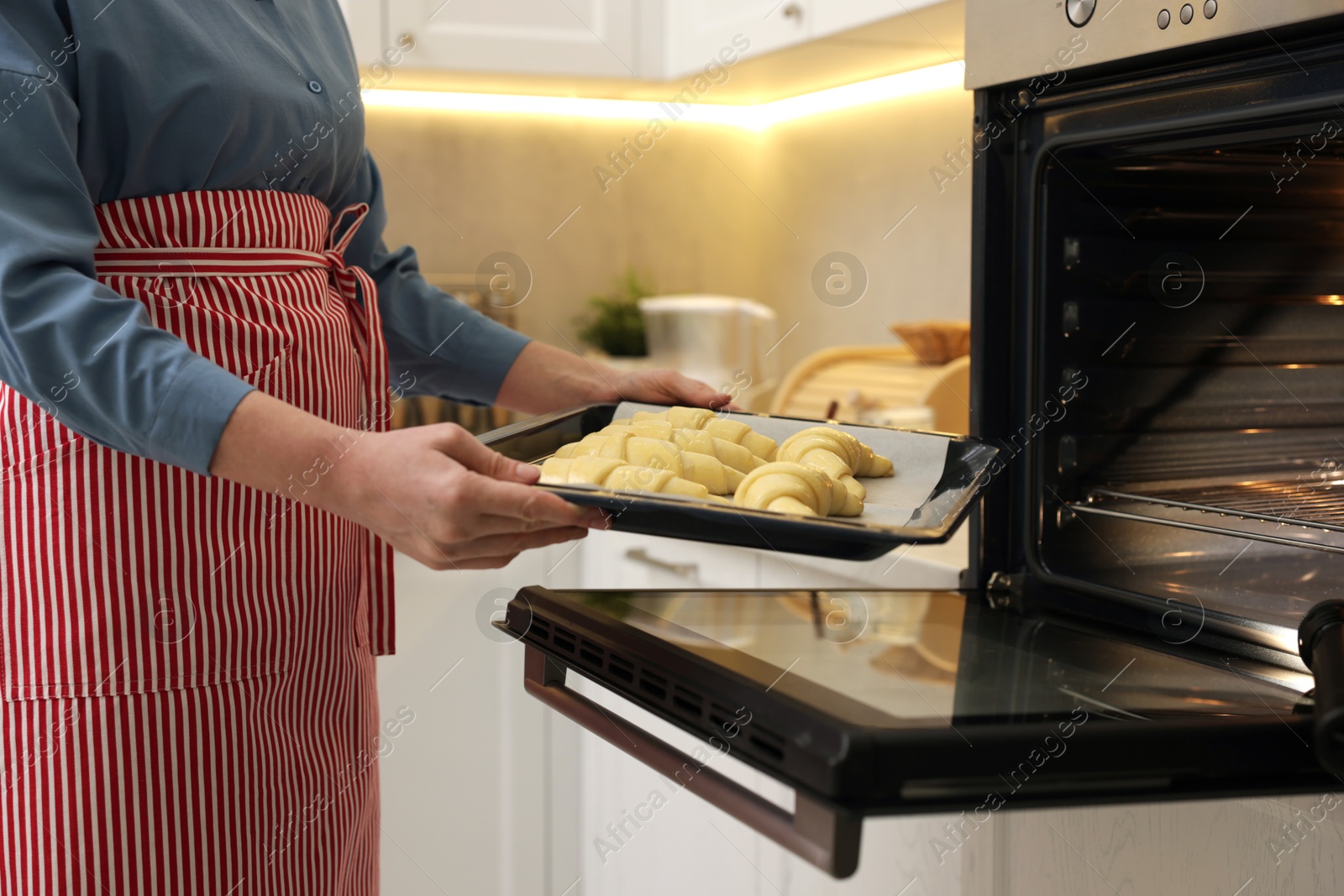 Photo of Woman putting baking sheet with raw croissants into oven indoors, closeup