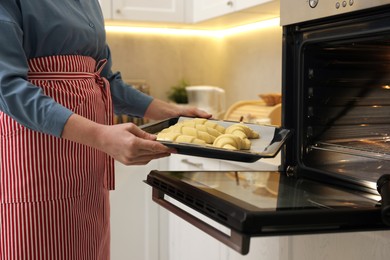 Photo of Woman putting baking sheet with raw croissants into oven indoors, closeup