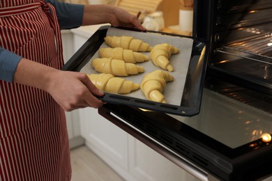 Woman putting baking sheet with raw croissants into oven indoors, closeup