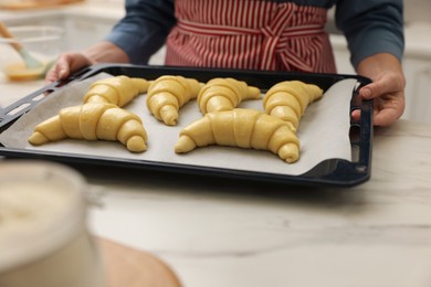 Photo of Woman taking baking sheet with raw croissants out of white marble table, closeup