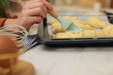 Photo of Woman brushing egg wash onto raw croissants at white table, closeup