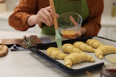 Woman brushing egg wash onto raw croissants at white marble table indoors, closeup