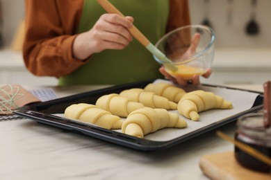 Photo of Woman brushing egg wash onto raw croissants at white marble table indoors, closeup