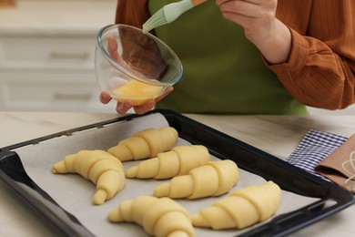 Photo of Woman brushing egg wash onto raw croissants at white marble table indoors, closeup