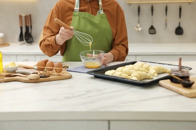 Photo of Woman whisking egg wash near raw croissants at white marble table in kitchen, closeup