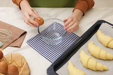 Photo of Woman breaking egg into bowl near raw croissants at white marble table, above view