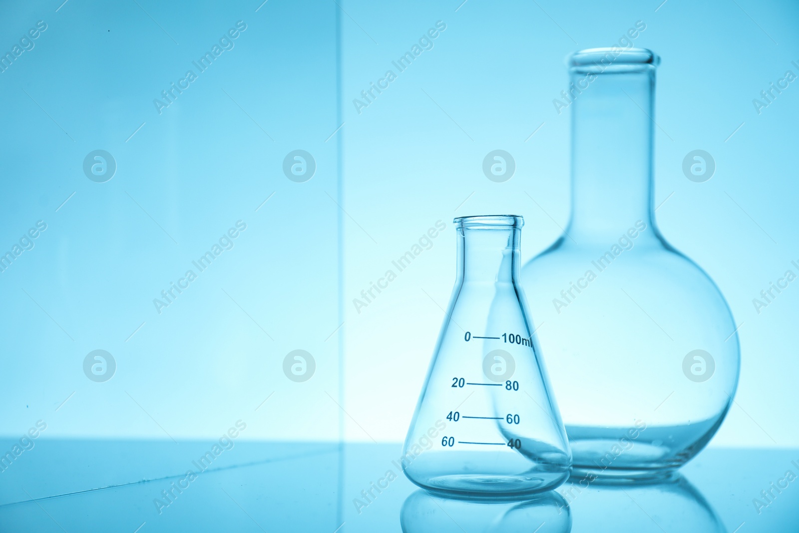 Photo of Empty flasks on mirror surface against light blue background, space for text. Laboratory equipment