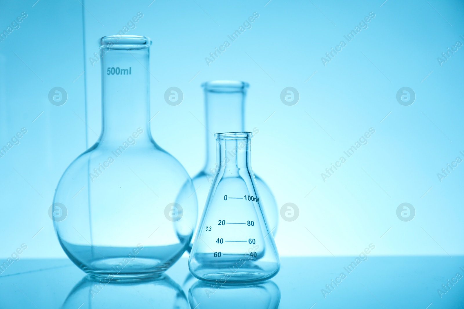 Photo of Empty flasks on mirror surface against light blue background, space for text. Laboratory equipment