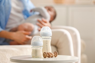 Photo of Mother holding her little baby indoors, focus on feeding bottles with milk and teether