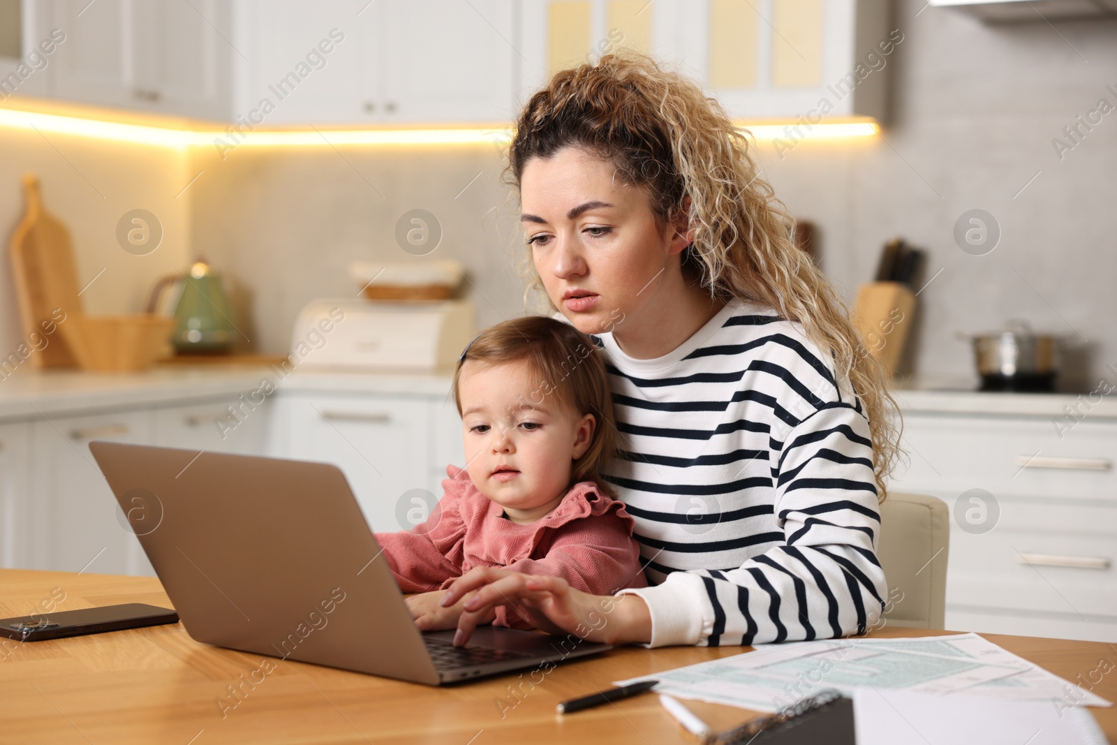 Photo of Single mother holding her daughter while working on laptop at wooden table in kitchen