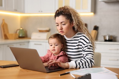 Single mother holding her daughter while working on laptop at wooden table in kitchen