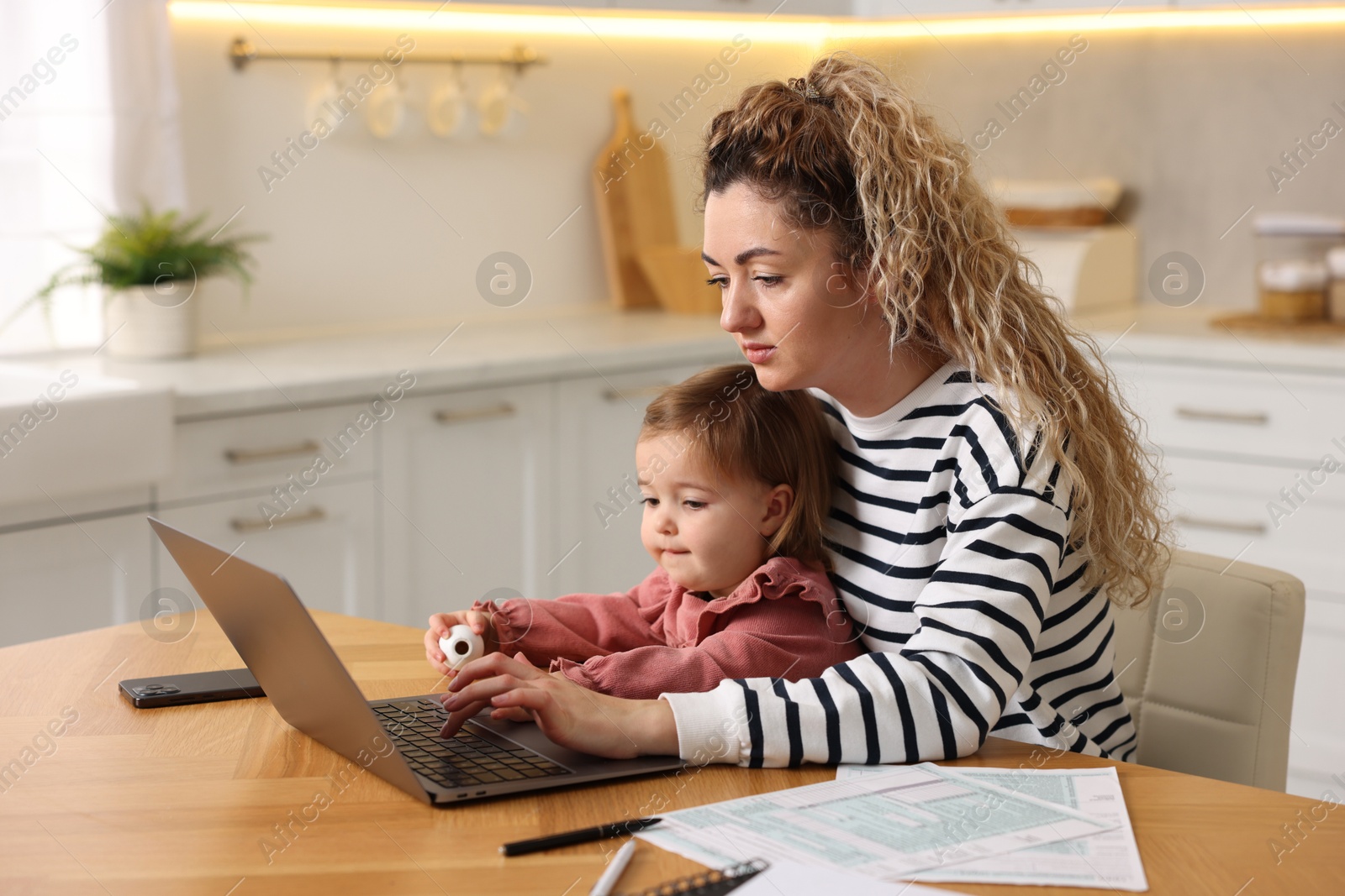 Photo of Single mother holding her daughter while working on laptop at wooden table in kitchen