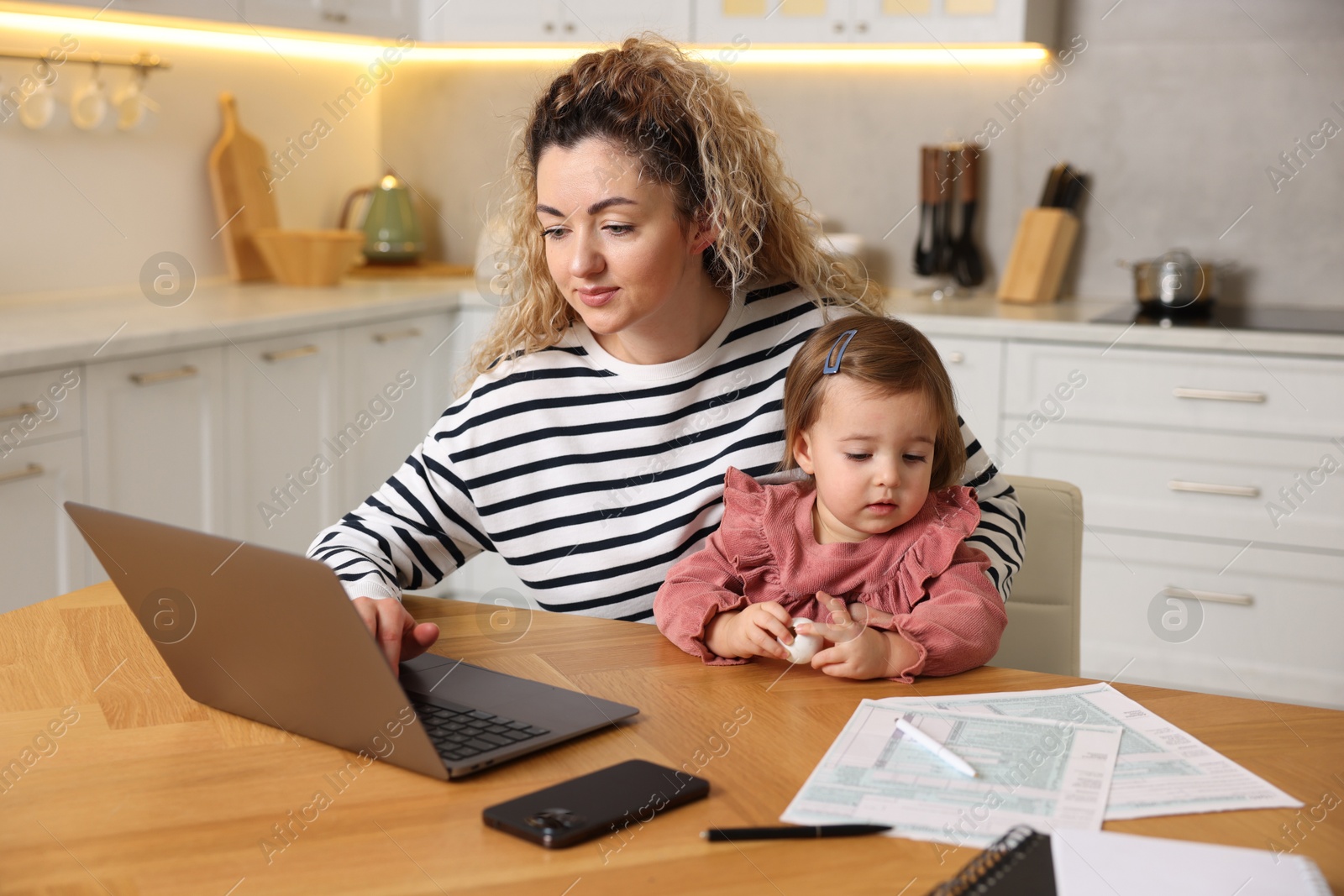 Photo of Single mother holding her daughter while working on laptop at wooden table in kitchen