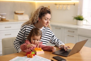 Photo of Single mother holding her daughter while working on laptop at wooden table in kitchen