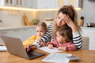 Photo of Single mother working on laptop and her daughters at table in kitchen