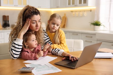 Photo of Single mother working on laptop and her daughters at table in kitchen