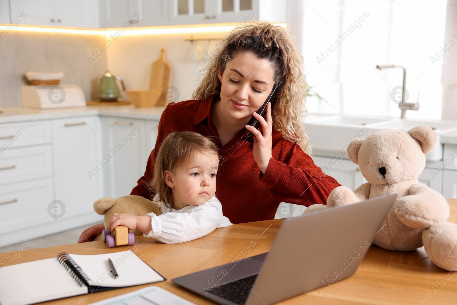 Photo of Work-family balance. Single mother holding her daughter while talking on smartphone at table in kitchen