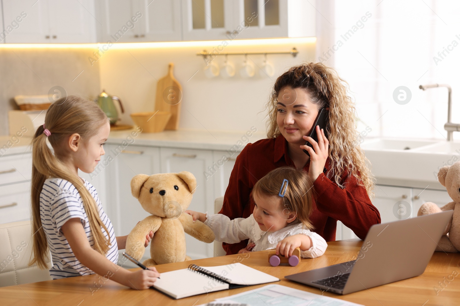 Photo of Work-family balance. Single mother talking on smartphone while her daughters playing at table in kitchen