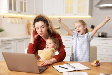 Photo of Single mother working with laptop while her daughters playing at table in kitchen