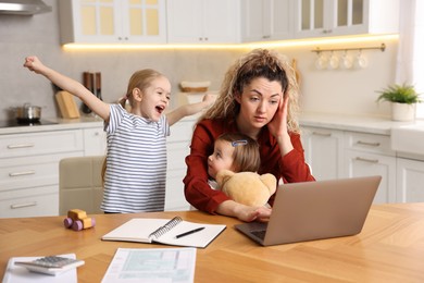 Photo of Single mother working with laptop while her daughters playing at table in kitchen