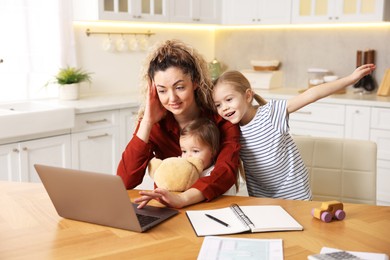 Photo of Single mother working with laptop while her daughters playing at table in kitchen