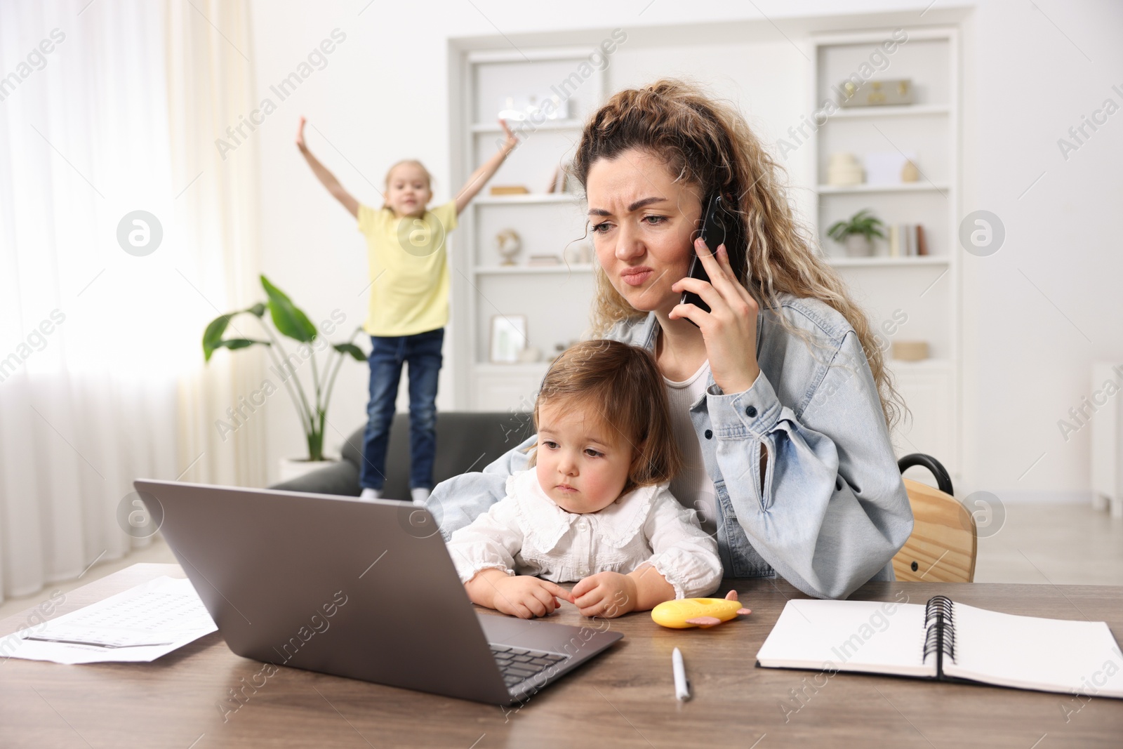 Photo of Work-family balance. Single mother holding daughter and talking on smartphone while her other child playing on sofa indoors, selective focus