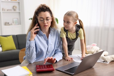 Photo of Work-family balance. Single mother using calculator while talking on smartphone and her daughter at table indoors