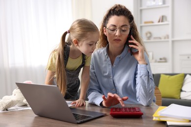 Photo of Work-family balance. Single mother using calculator while talking on smartphone and her daughter at table indoors