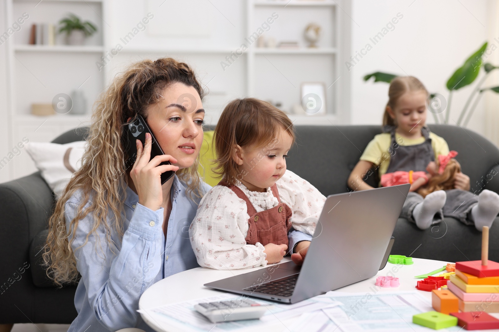 Photo of Work-family balance. Single mother holding daughter and talking on smartphone while her other child playing on sofa indoors, selective focus