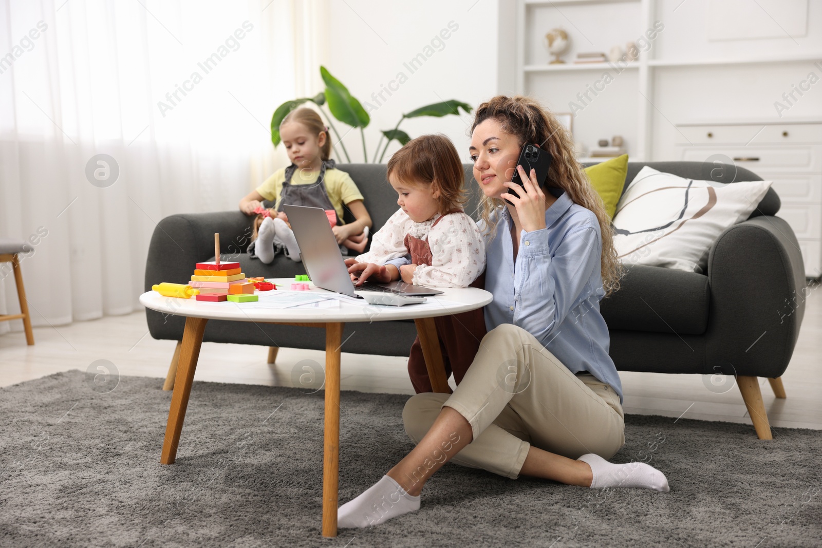 Photo of Single mother talking on smartphone and working with laptop while her daughters playing indoors