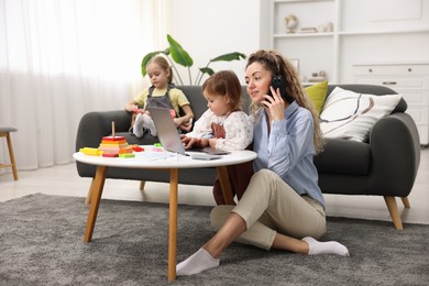 Single mother talking on smartphone and working with laptop while her daughters playing indoors